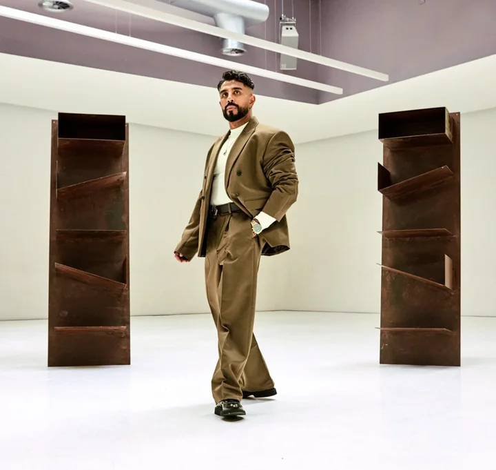 A man in a loose brown suit walking through a white gallery between two tall rusted steel sculptures