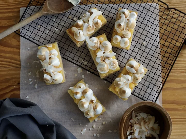 Six lemon meringue slices topped with toasted coconut flakes on baking paper and a wire rack, with a wooden spoon and a bowl of coconut alongside